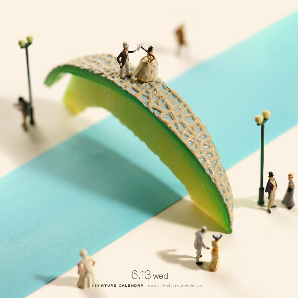 Miniature scene by Tatsuya Tanaka depicting tiny figurines crossing a bridge made from a slice of melon rind, with street lamps and pedestrians on a blue and white striped surface.