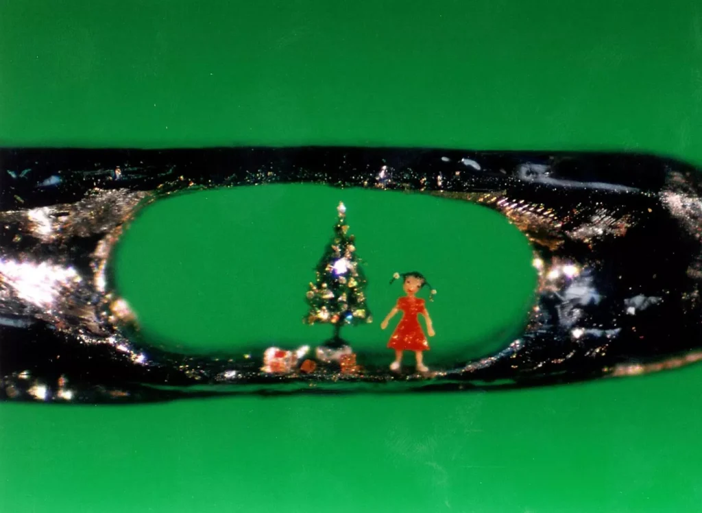 Micro-sculpture by Willard Wigan depicting a girl in a red dress standing beside a decorated Christmas tree with presents, all inside the eye of a sewing needle against a green background.
