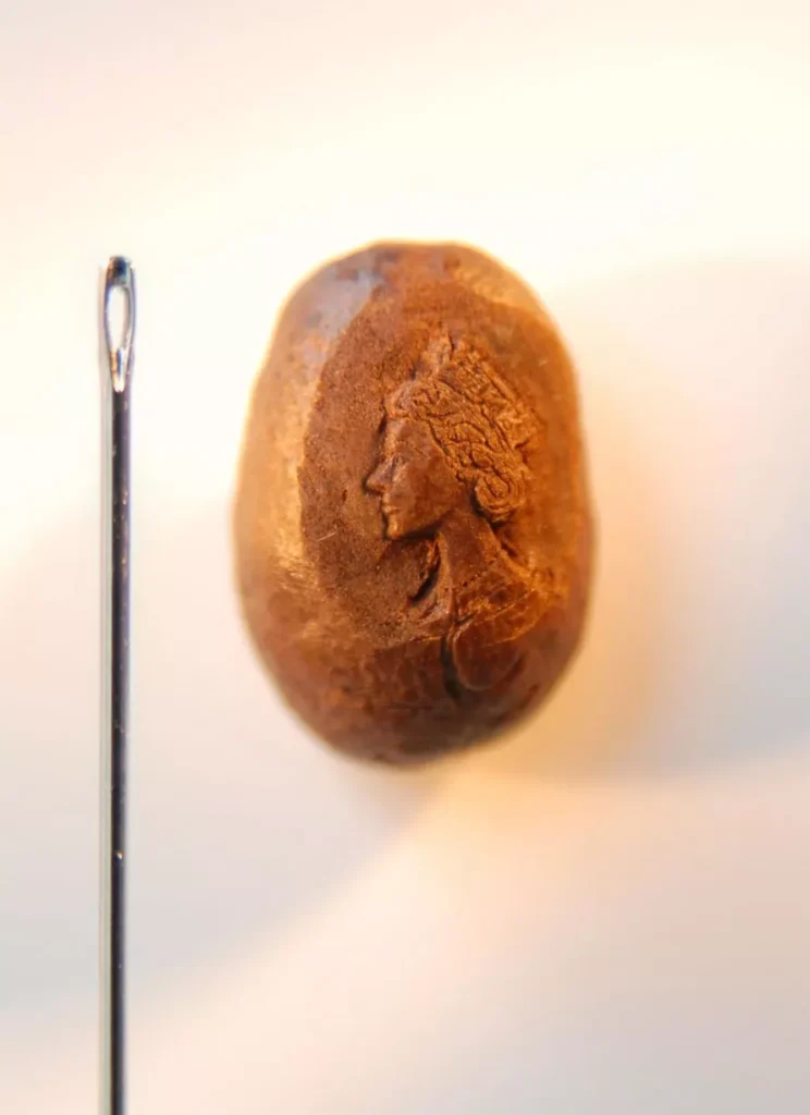 Micro-sculpture by Willard Wigan featuring a detailed portrait of Queen Elizabeth II carved into a single coffee bean, shown next to the eye of a sewing needle for scale.