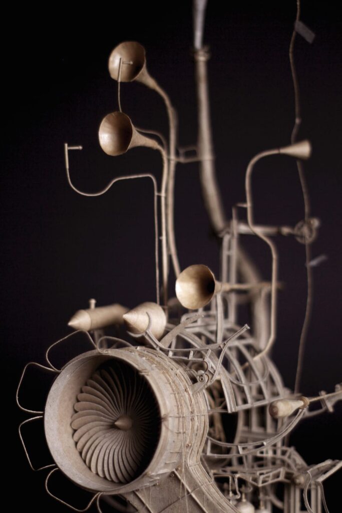 Close-up of Daniel Agdag’s cardboard sculpture The Pilot, showcasing intricate steampunk-inspired details including a large turbine engine, curved pipes, and horn-like structures, all meticulously crafted from sliced cardboard and PVA glue against a dark background.