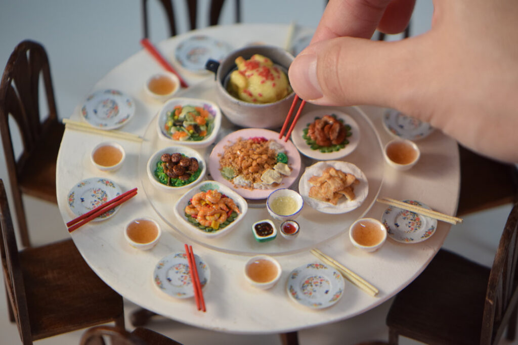 Miniature Chinese banquet scene by Pui Wan Lim, featuring a round dining table set with tiny plates, bowls, chopsticks, and lifelike clay dishes, with a hand using chopsticks to pick up food.