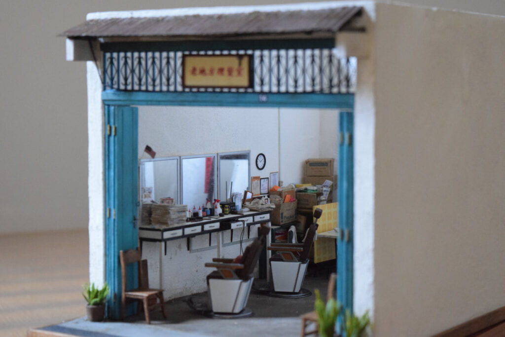 Miniature model by Pui Wan Lim of a vintage Malaysian barbershop, featuring blue-framed open doors, barber chairs, mirrors, and detailed interior props like newspapers, boxes, and hairdressing tools.
