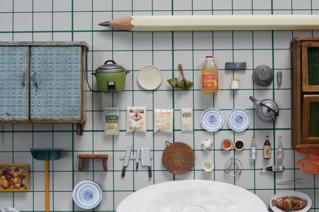 Miniature kitchen scene by Pui Wan Lim featuring an overhead view of dollhouse-scale cooking utensils, food items, condiments, plates, and furniture arranged neatly on a tiled surface with a pencil for scale.