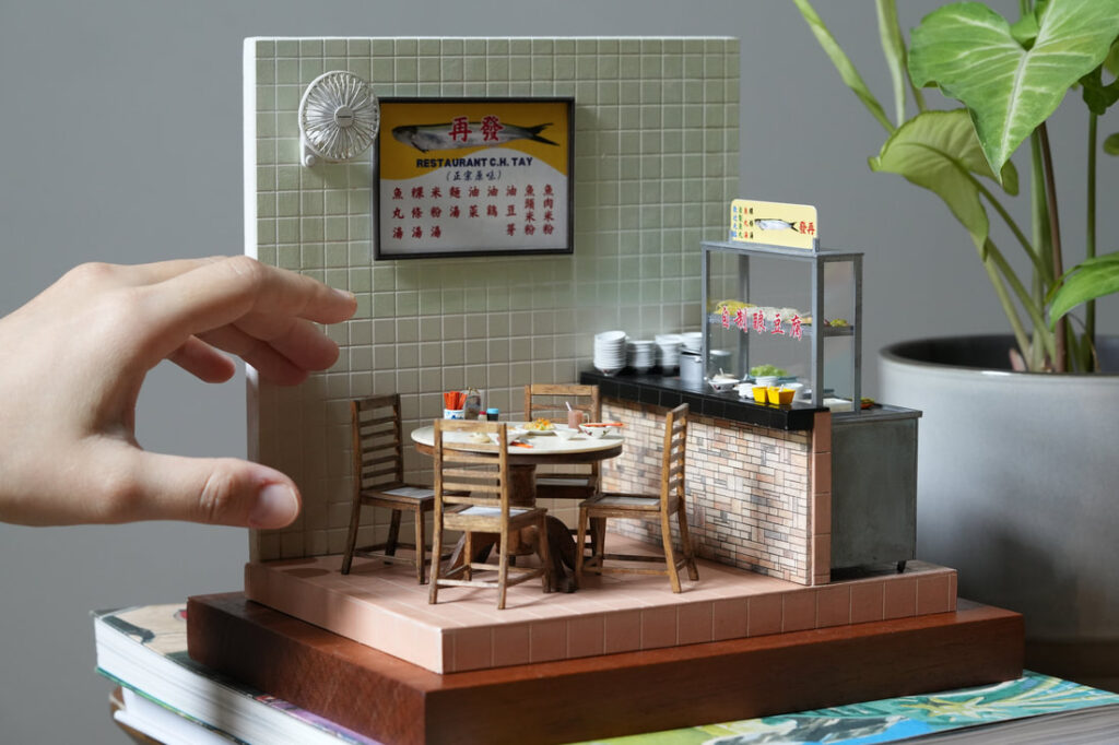 Miniature diorama by Pui Wan Lim of a Malaysian restaurant scene, featuring a tiled wall with signage, a round dining table set with dishes, wooden chairs, and a food stall counter, with a hand shown for scale.